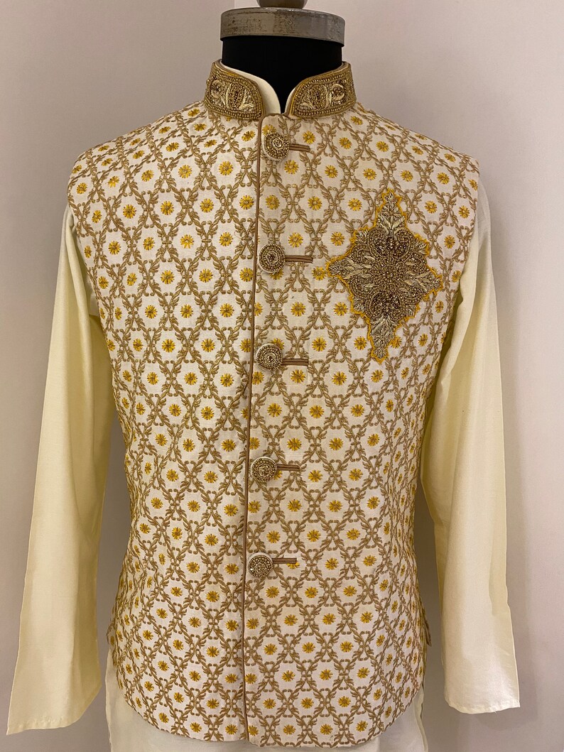 May include: A white waistcoat with a gold and yellow geometric pattern and a gold embroidered design on the front. The waistcoat has a mandarin collar and a button closure.