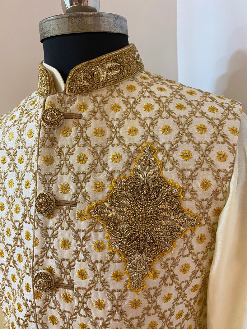 May include: A cream-coloured waistcoat with gold embroidery and a beaded design. The waistcoat has a mandarin collar and a button closure.