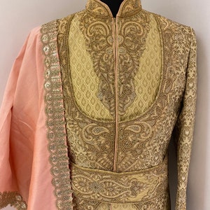 May include: A cream-colored, intricately embroidered jacket with gold thread detailing. The jacket features a high collar and a matching belt. A light pink shawl with gold sequin trim is draped over the shoulder. The garment is displayed on a mannequin.