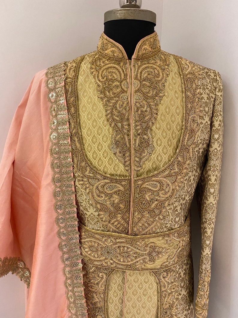 May include: A cream-colored, intricately embroidered jacket with gold thread detailing. The jacket features a high collar and a matching belt. A light pink shawl with gold sequin trim is draped over the shoulder. The garment is displayed on a mannequin.