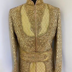 May include: A gold-colored, intricately embroidered sherwani suit. The long-sleeved jacket features ornate patterns, a mandarin collar, and a decorative belt. The suit is displayed on a mannequin, showcasing the detailed craftsmanship and design.