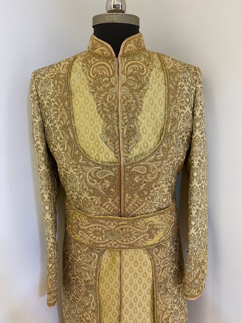 May include: A gold-colored, intricately embroidered sherwani suit. The long-sleeved jacket features ornate patterns, a mandarin collar, and a decorative belt. The suit is displayed on a mannequin, showcasing the detailed craftsmanship and design.