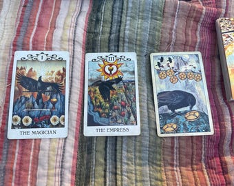 May include: Three tarot cards are displayed, featuring illustrations of ravens and symbolic imagery. The cards are labeled "The Magician," "The Empress," and "VIII." A deck of cards is visible in the background, all resting on a striped and patterned surface.