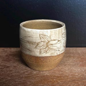 Może przedstawiać: Mały ceramiczny kubek z dwukolorowym wykończeniem. Górna część pokryta jest gładkim, kremowobiałym szkliwem z wygrawerowanym ciemnobrązowym rysunkiem kwiatu narcyza. Dolna część wykonana jest z surowej, brązowej gliny o wyraźnej fakturze.