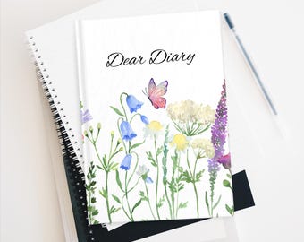 May include: A white journal with the words "Dear Diary" in script, adorned with watercolor illustrations of wildflowers and a butterfly. A pen rests beside the journal, which is stacked on top of other notebooks.