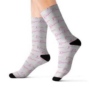 Puede incluir: Calcetines blancos con una escritura rosa repetida que dice "Dance" y "Grace". Los calcetines tienen talones y dedos negros. Estos calcetines son un accesorio divertido para bailarines o cualquier persona que ame bailar.