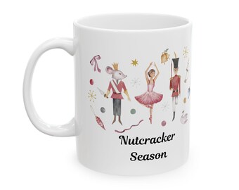 May include: White ceramic mug featuring a Nutcracker Season design. The design includes illustrations of a mouse, a ballerina, and a soldier in red and gold uniforms, along with other festive elements. The mug has a curved handle.