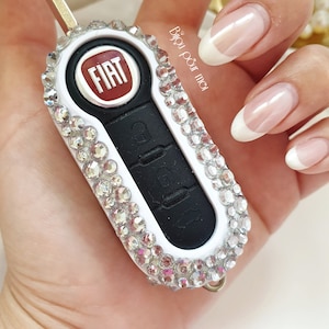 May include: A white car key with a black button panel and a red FIAT logo. The key is covered in clear rhinestones.