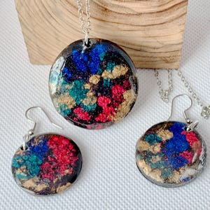 May include: A jewelry set featuring a pendant necklace and matching earrings. The round pendants and earrings have a black background with splashes of blue, teal, red, and gold. The necklace and earrings have silver-colored chains and hooks.