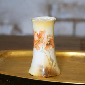 May include: A cylindrical porcelain vase with a flared top and base, featuring a hand-painted design of orange poppy flowers on a cream-colored background. The vase has a soft yellow gradient at the top rim and a subtle brown shading at the base.