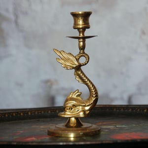 May include: A brass candlestick holder with a dolphin design. The candlestick features a detailed dolphin with a curved tail and a fin-like element. The holder is designed to hold a single candle and has a round base.