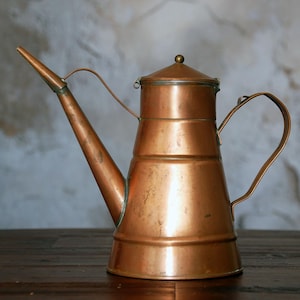 May include: A copper-colored, antique oil can with a long spout and a curved handle. The can has a lid with a small brass knob and is made of copper. The design is vintage and the can is likely used for pouring liquids.
