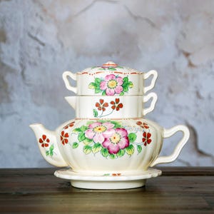 May include: A cream-colored ceramic tea set with a teapot, creamer, and sugar bowl stacked on a saucer. The set is decorated with pink and green floral designs and red accents. The teapot has a curved spout and handle.