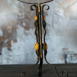 May include: A vintage-style vanity mirror with a bronze-colored metal frame and stand. The stand features decorative floral accents in yellow and green. The mirror is round and has a decorative edge.