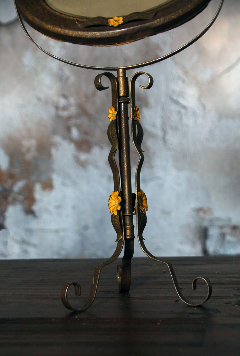 May include: A vintage-style vanity mirror with a bronze-colored metal frame and stand. The stand features decorative floral accents in yellow and green. The mirror is round and has a decorative edge.