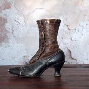 May include: An antique black and brown leather boot with a pointed toe and a small heel. The boot has a button closure on the side and is likely from the Victorian era. The leather shows signs of wear and age, adding to its vintage charm.