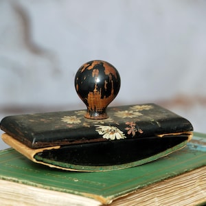 May include: An antique inkwell with a black, floral-decorated lid and a round, dark wooden handle. The inkwell rests on a green book with aged pages, suggesting a vintage aesthetic. The inkwell's design includes white and pink floral accents.