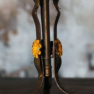 May include: Close-up of a decorative metal stand with a floral design. The stand has a dark bronze finish with yellow flowers and green leaves. The stand's legs curve gracefully, adding to its ornate appearance.