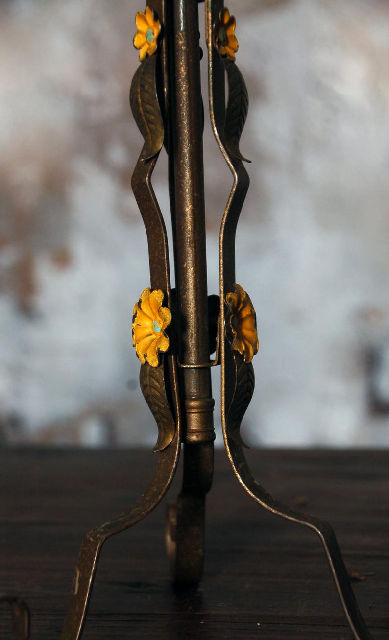 May include: Close-up of a decorative metal stand with a floral design. The stand has a dark bronze finish with yellow flowers and green leaves. The stand's legs curve gracefully, adding to its ornate appearance.