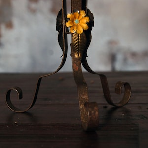May include: Close-up of a decorative metal stand with a floral accent. The stand has a distressed, antique finish with a yellow flower and turquoise center. The legs have a scrolled design.