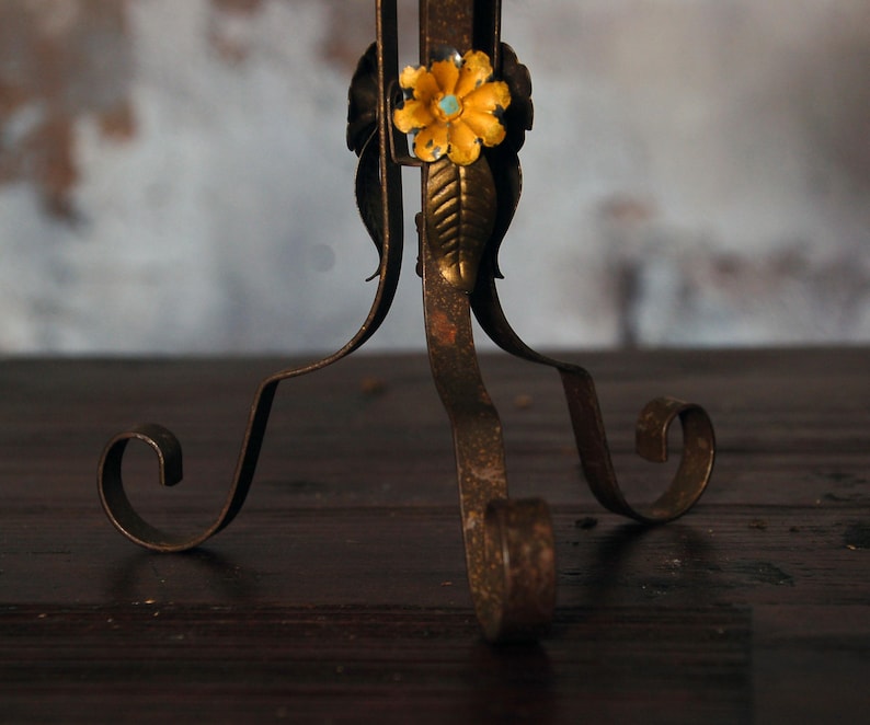 May include: Close-up of a decorative metal stand with a floral accent. The stand has a distressed, antique finish with a yellow flower and turquoise center. The legs have a scrolled design.