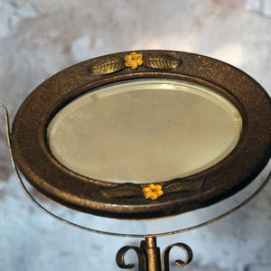 May include: Oval vanity mirror with a bronze-colored frame and stand. The frame is decorated with small yellow flowers and leaf accents. The mirror is supported by a curved metal stand.