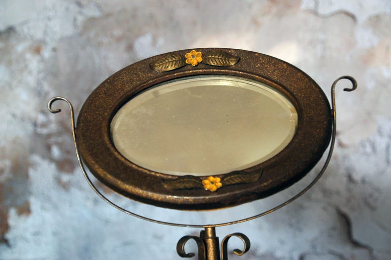 May include: Oval vanity mirror with a bronze-colored frame and stand. The frame is decorated with small yellow flowers and leaf accents. The mirror is supported by a curved metal stand.