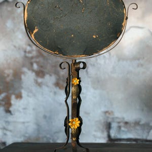 May include: An antique, round vanity mirror with a dark, textured surface. The mirror is supported by a decorative, wrought-iron stand with floral accents. The stand has a curved base and is finished in a rustic, aged bronze color.