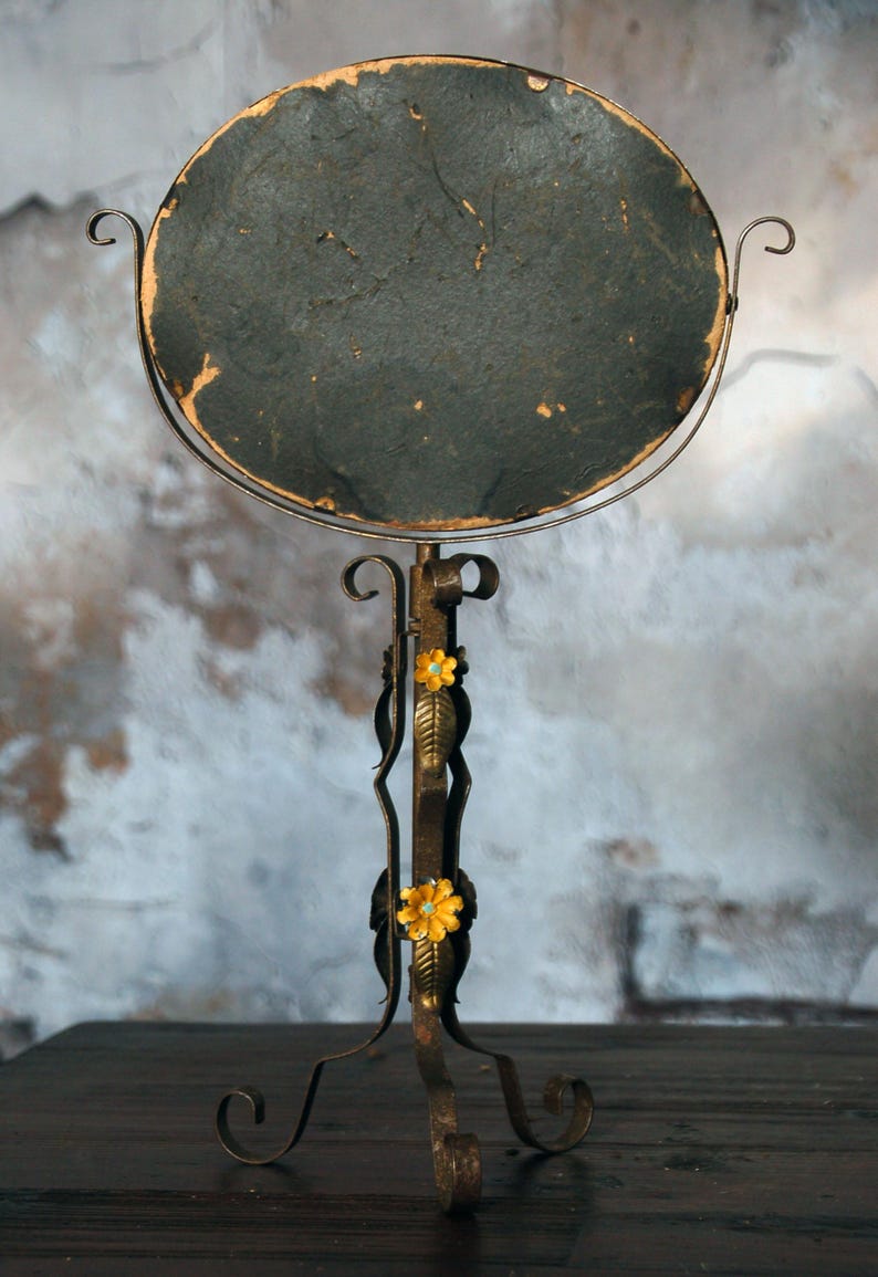May include: An antique, round vanity mirror with a dark, textured surface. The mirror is supported by a decorative, wrought-iron stand with floral accents. The stand has a curved base and is finished in a rustic, aged bronze color.