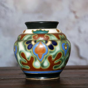 May include: A small ceramic vase with a bulbous shape, featuring a black rim and base. The body is decorated with a repeating pattern of stylized floral and leaf motifs in shades of green, burnt orange, deep blue, and white with black outlines.