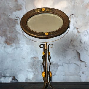 May include: An antique vanity mirror with an oval-shaped, bronze-colored frame. The frame is adorned with yellow floral accents and rests on a decorative stand. The stand has a curved design with yellow floral details and a three-legged base.