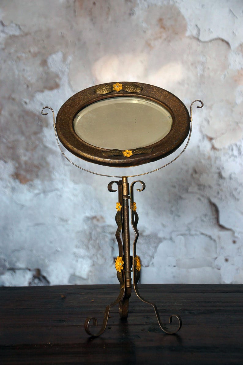 May include: An antique vanity mirror with an oval-shaped, bronze-colored frame. The frame is adorned with yellow floral accents and rests on a decorative stand. The stand has a curved design with yellow floral details and a three-legged base.