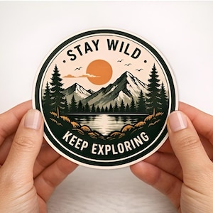 May include: A round sticker with a nature scene featuring mountains, a lake, and trees under a setting sun. The sticker has the words "STAY WILD" and "KEEP EXPLORING" in a circular design, with a black border and a white background.