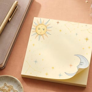 May include: A stack of celestial-themed sticky notes. The top note features a sun with a face and a crescent moon with a sleeping face. The notes are decorated with stars and constellations in yellow, blue, and orange on a cream background. A gold pen and notebook are in the background.