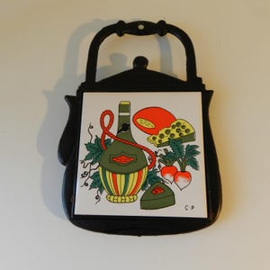 May include: Black cast iron trivet with a handle, featuring a ceramic tile with a wine and cheese design. The tile depicts a green bottle, cheese, radishes, and a red rope. The trivet is designed to protect surfaces from hot items.