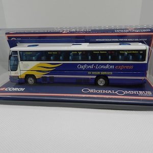 Peut inclure: Un modèle de bus blanc et bleu avec les mots "Oxford London express" et "24 hour service" sur le côté. Le bus est dans une vitrine en plastique transparent avec un logo Corgi au fond.