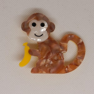 May include: A monkey-shaped brooch with a brown and tan marbled pattern. The monkey holds a yellow banana. The brooch has a white face with black eyes and a smiling mouth. The tail curls around the body.