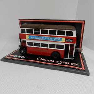 May include: A red double-decker bus model with a brown roof and white accents. The bus has a blue and white advertisement that reads "Buy Savings Certificates". The model is displayed on a black base with the words "The Original Omnibus Company" printed on it.