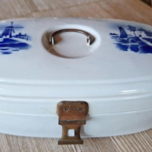 Könnte beinhalten: Ovale Brotbox aus weißer Keramik mit blauen Windmühlen- und Seelandschaftsdesigns auf dem Deckel. Die Box hat einen Metallgriff und einen Metallverschluss. Ein Vintage-Küchenartikel.