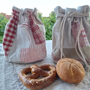 Könnte beinhalten: Zwei Beutel mit Kordelzug aus beigem Stoff mit rot-weißem Patchwork-Muster, einschließlich Vichy-Karos und Streifen. Die Beutel haben dicke, cremefarbene Kordeln. Eine Salzbrezel und ein rundes Brötchen liegen vor den Beuteln auf dem Untergrund.