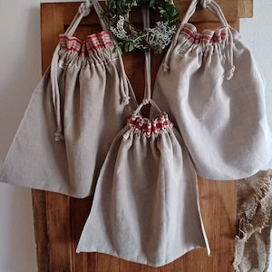 Könnte beinhalten: Drei beigefarbene Leinensäckchen mit rotem und weißem Streifenbesatz hängen an einer Holztür. Die Säcke sind leer und bereit zum Befüllen.