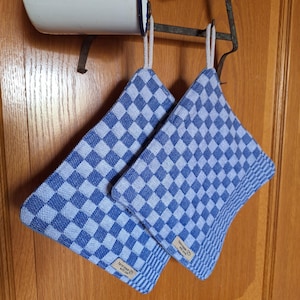 Könnte beinhalten: Zwei blau-weiß karierte Topflappen hängen an einem Metallgestell. Die Topflappen haben ein quadratisches Muster und sind aus Stoff. Die Topflappen hängen an einer Holztür.