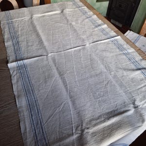 Könnte beinhalten: Eine weiße Tischdecke aus Leinen mit blau gestreiften Rändern. Das Tuch liegt auf einem Holztisch und zeigt die Textur des Stoffes. Die blauen Streifen verlaufen entlang der Ränder des rechteckigen Tuches.