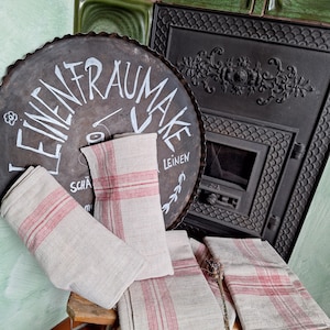 Könnte beinhalten: Mehrere Leinentücher mit roten Streifen sind auf einem Holzschemel drapiert. Die Tücher sind gefaltet und in einer rustikalen, Vintage-Ästhetik angeordnet. Im Hintergrund befindet sich ein großes, rundes Metallschild mit weißer Schrift.