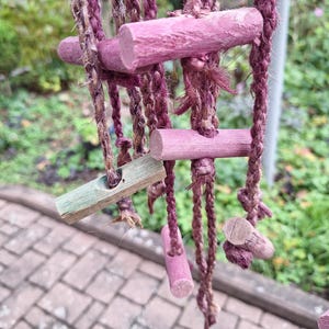 Könnte beinhalten: Nahaufnahme eines hängenden Pflanzenhalters mit Leiterdesign. Der Pflanzenhalter besteht aus geflochtenem Seil und verfügt über rosa Holzstäbe. Das Seil hat eine Mischung aus Braun- und Lilatönen, mit einem grünen Dübel unten.