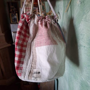 Könnte beinhalten: Eine weiße, rote und blaue Patchwork-Kordeltasche mit einem handgefertigten Etikett. Die Tasche ist aus verschiedenen Stoffen gefertigt, darunter Gingham und Leinen.