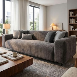 May include: A large, modern grey sofa with several grey scatter cushions. The sofa has a low profile and rounded arms. A wooden coffee table with books and a mug sits in front. The room has large windows with sheer curtains.