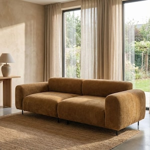 May include: A modern, two-cushion sofa in a warm, golden-brown color. The sofa has rounded arms and sits on black metal legs. A neutral-toned rug is beneath the sofa, and sheer curtains frame large windows. A small table with a lamp is visible.