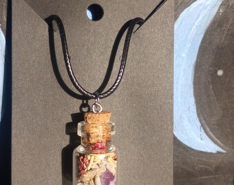 May include: A glass bottle necklace with a cork stopper filled with dried flowers, herbs, and crystals. The necklace is on a black cord and is hanging from a black card.