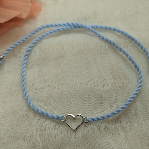 May include: A light blue friendship bracelet with a silver heart charm. The bracelet has two tassels on the ends.